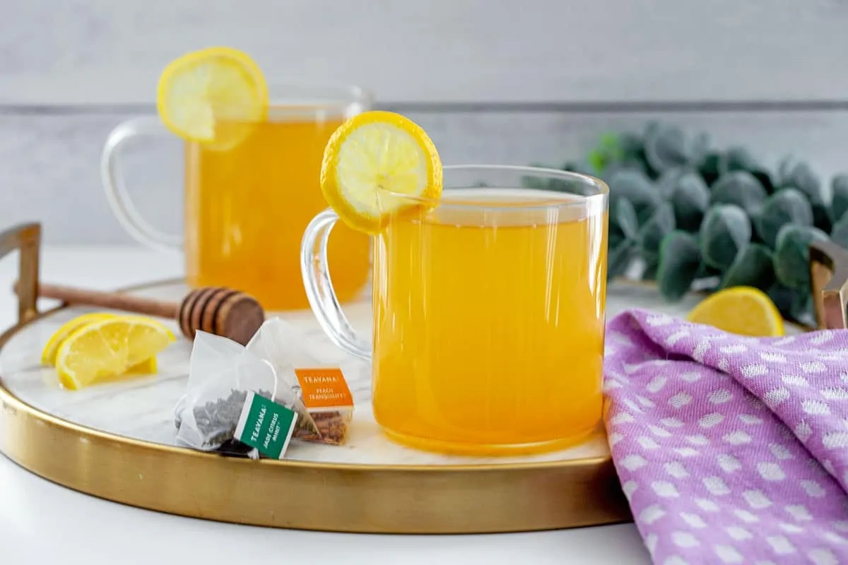 Khung cảnh hai cốc Honey Citrus Mint Tea đã hoàn thành, với lát chanh và túi trà đặt ở mặt bàn.